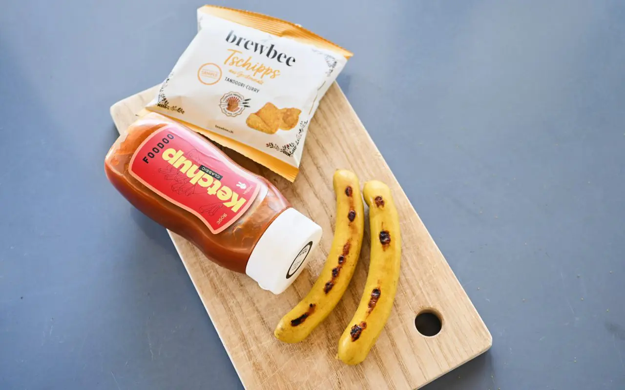 Holzbrett mit einer Flasche Ketschup, Chips und zwei Curry Würsten