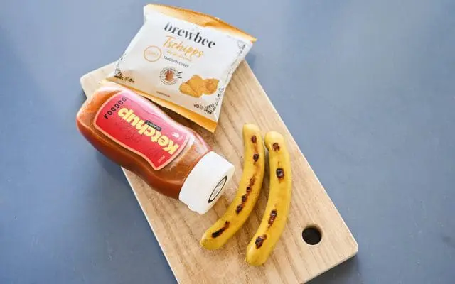 Holzbrett mit einer Flasche Ketschup, Chips und zwei Curry Würsten