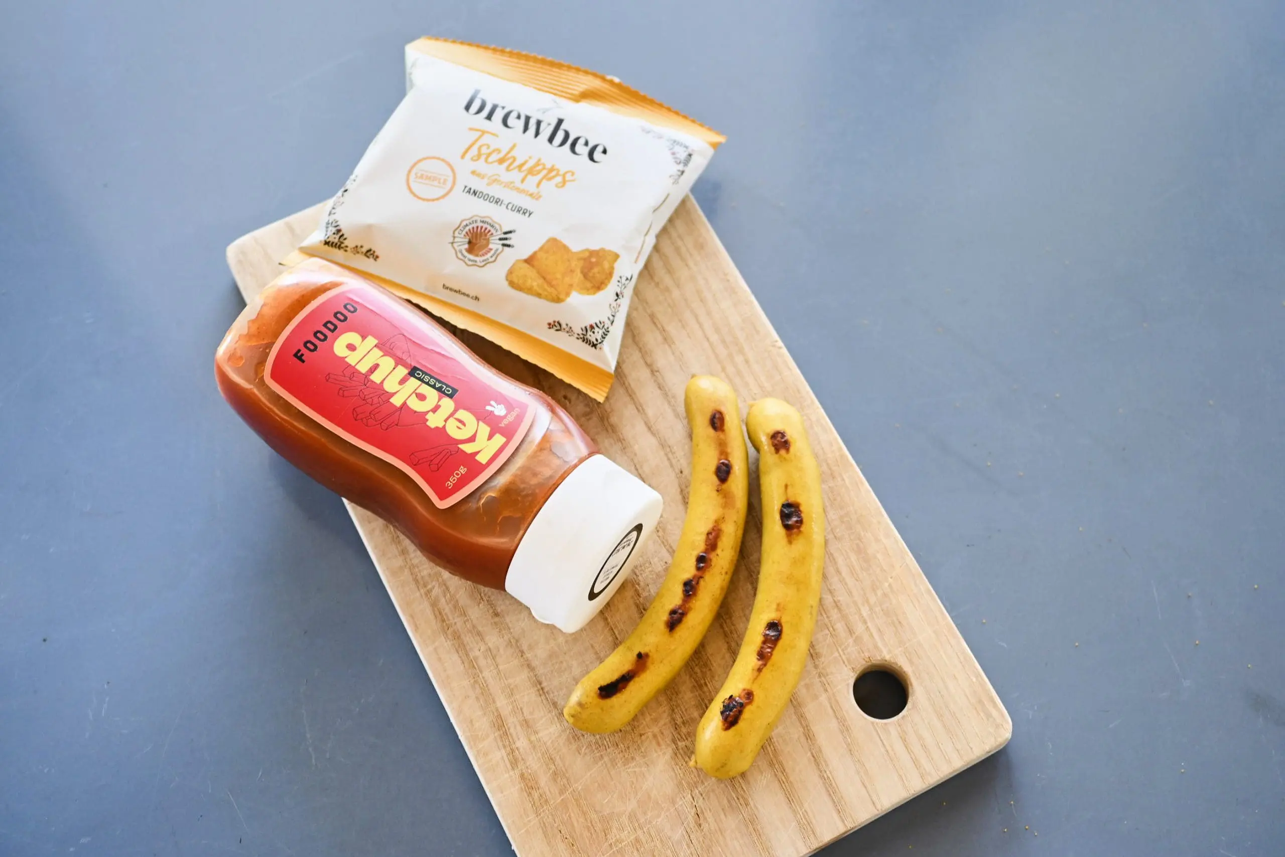 Holzbrett mit einer Flasche Ketschup, Chips und zwei Curry Würsten